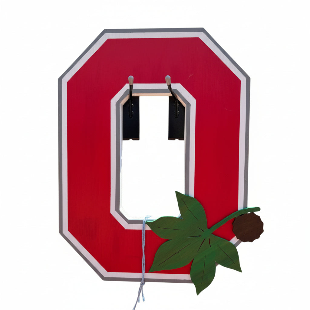 A large red and white cutout letter 'O' mounted on a black metal fence, with a green maple leaf decoration hanging in front. A street scene with parked cars and buildings is visible behind the sign.
