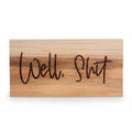 Wooden sign with 'Well, Shit' engraved on it, surrounded by greenery.