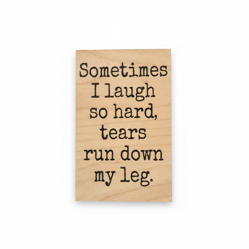 A rectangular light wood sign with black engraved text that reads, 'Sometimes I laugh so hard, tears run down my leg.' The sign is photographed against a dark background.